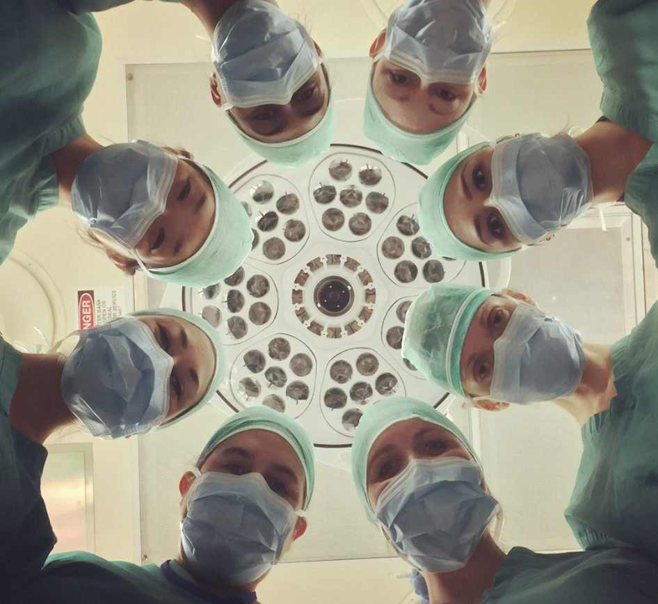 A surgical care team looking upward together beneath an operating room light, viewed from below in a circular formation.